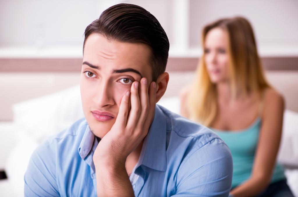 Man looking frustrated or disappointed, perhaps after an intimate moment, partner looks concerned or disappointed in the background. Sad mood.